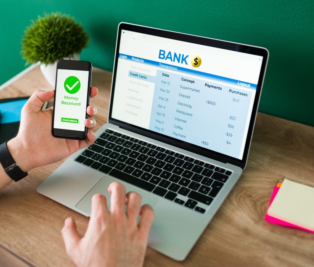 Wire transfer. Young man using his smartphone and laptop to do a banking transaction and some payments